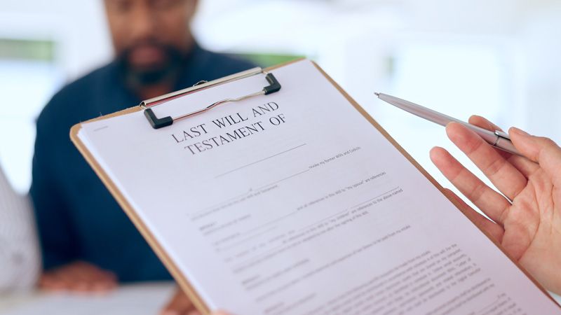 A woman holds a clipboard that reads "Last Will And Testament Of" in the foreground. In the background, a man's face is half visible and blurred out.