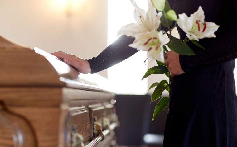 A person places their hand on a casket while holding flowers.